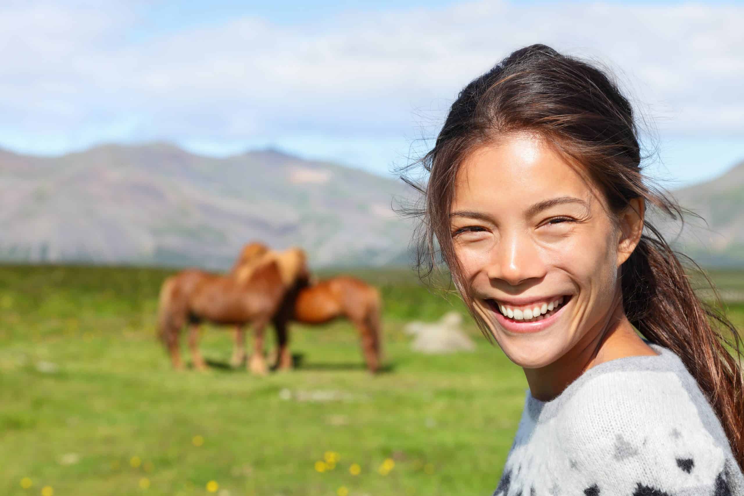 Woman on Iceland smiling with Icelandic horses. Portrait of happy multicultural girl wearing Icelandic sweater standing outdoors in nature field in front of horse.