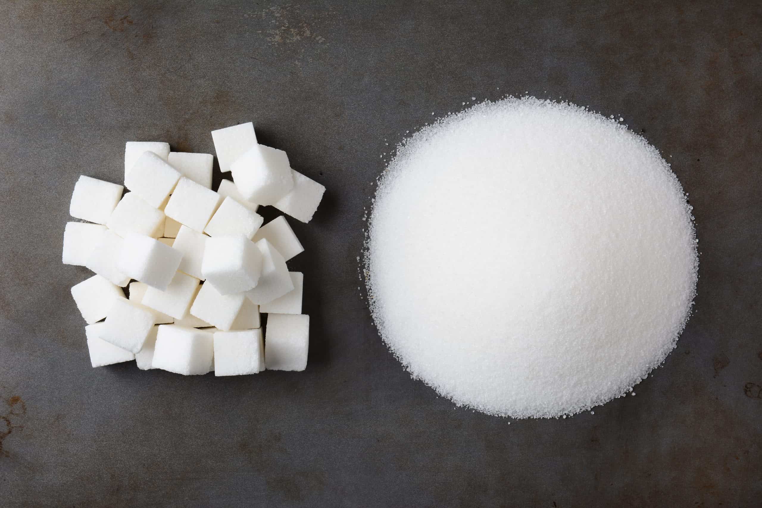 Overhead view of a pile of granulated white sugar and a mound of sugar cubes, on a used baking sheet.