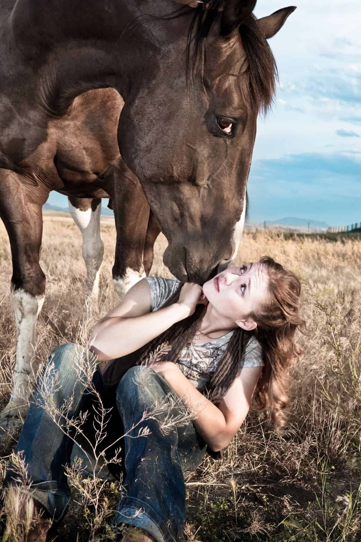 Brunette girl sitting on ground with horse above givin gkiss