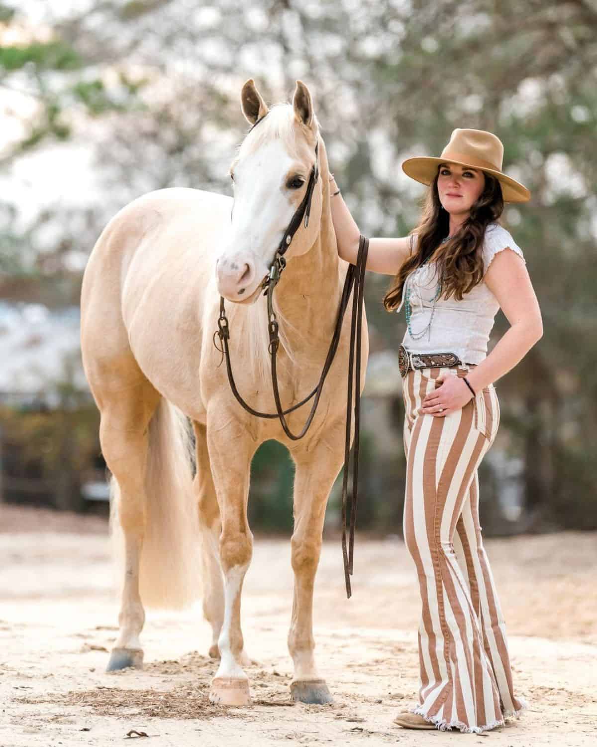 A young woman stands next to a light-brown horse.
