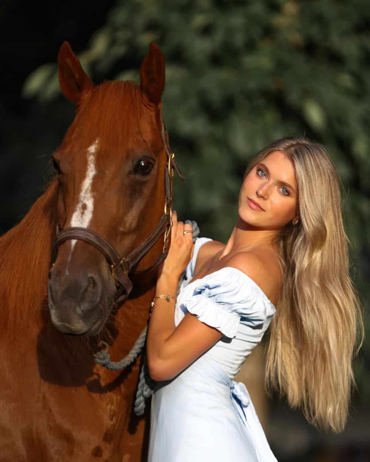 A young woman stands near a brown horse.