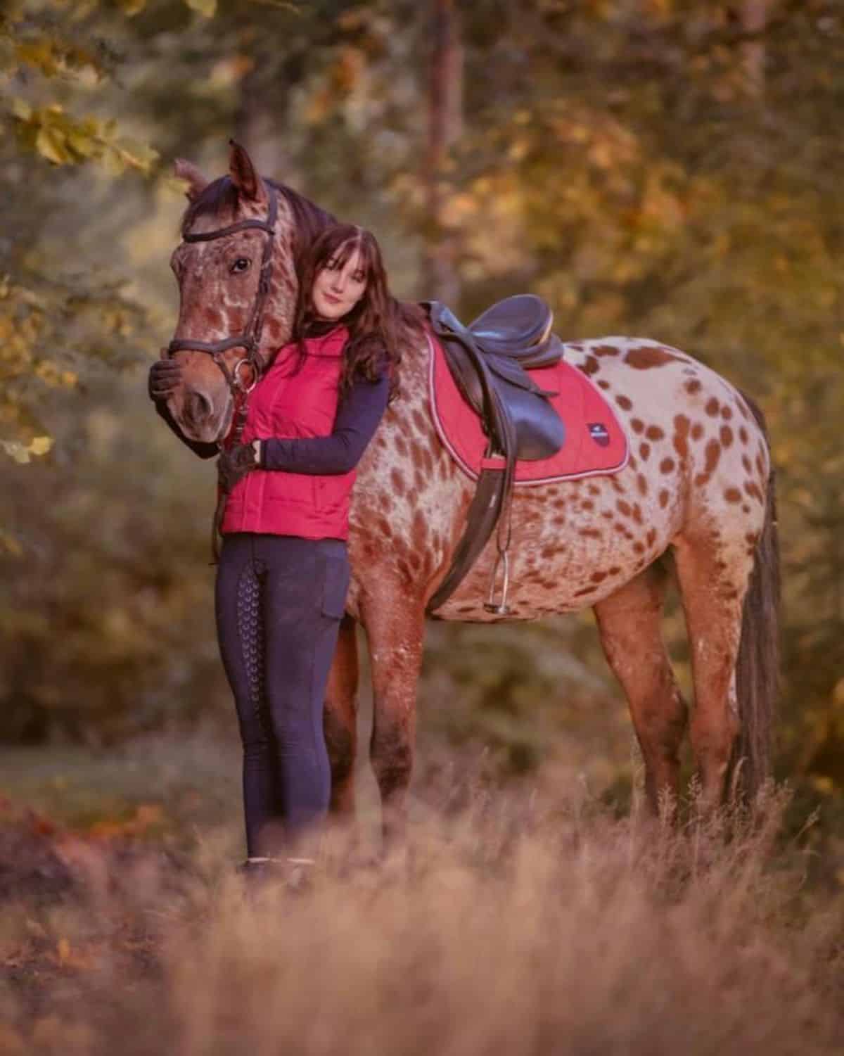 A young woman stands near brown spotted horse.