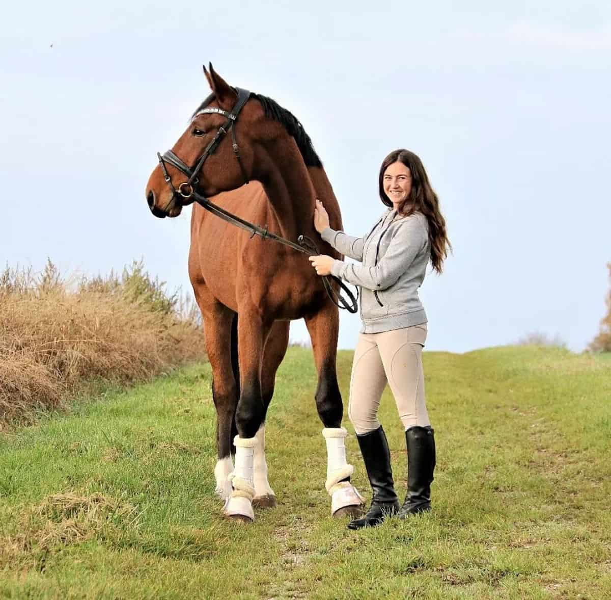 A young woman stands near a brown horse.