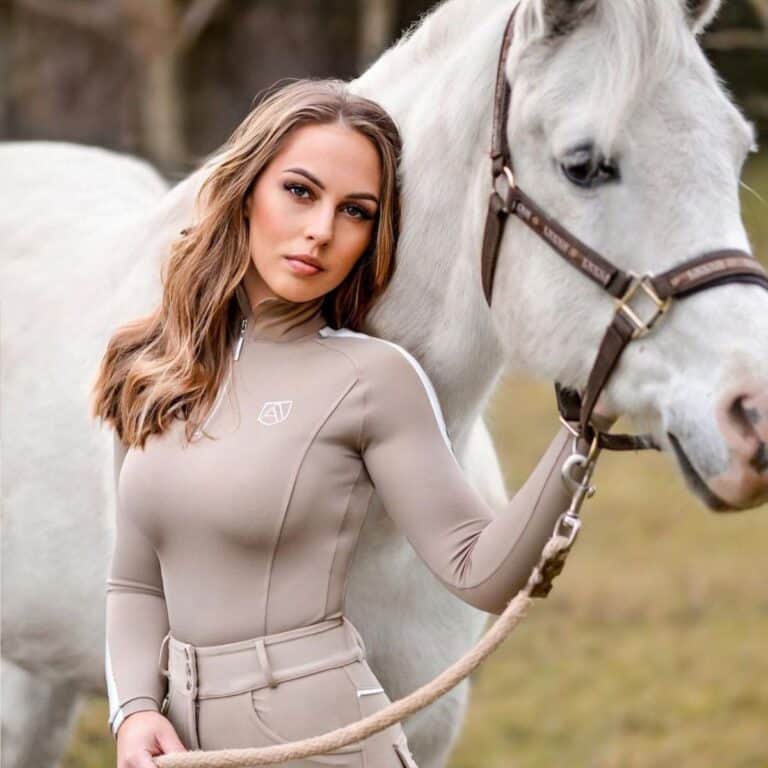 A young woman in an equestrian outfit stands next to a white horse.