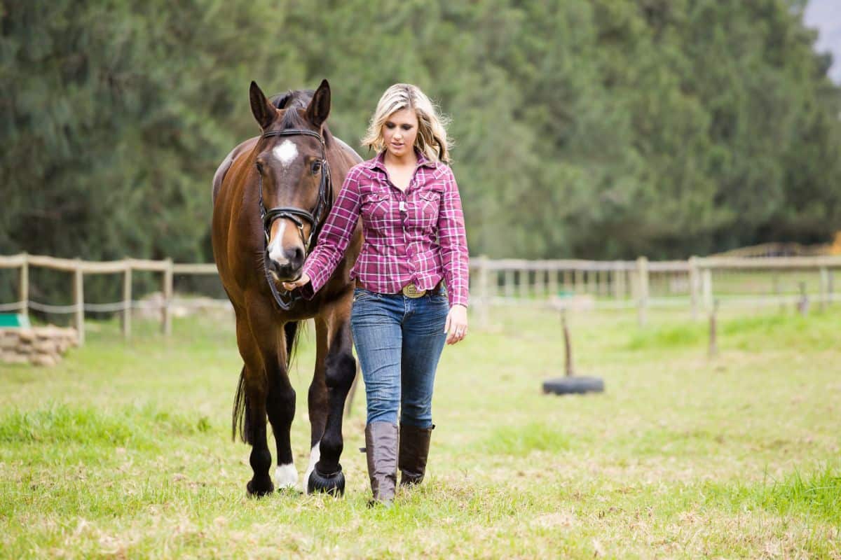 A young woman leading a brown horse on a ranch.