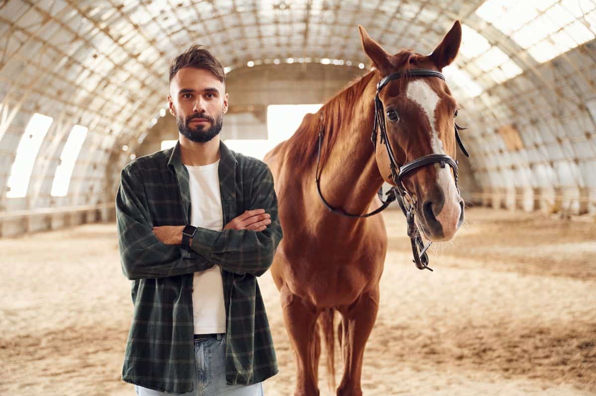 A young man stands near a brown horse.