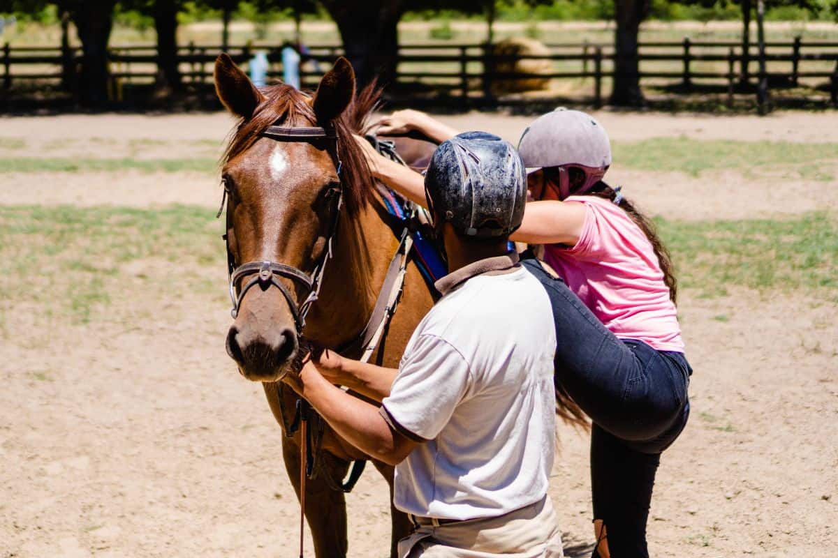 A young girl taking a horseback riding lesson while a horsetrainer holding a horse by a halter.