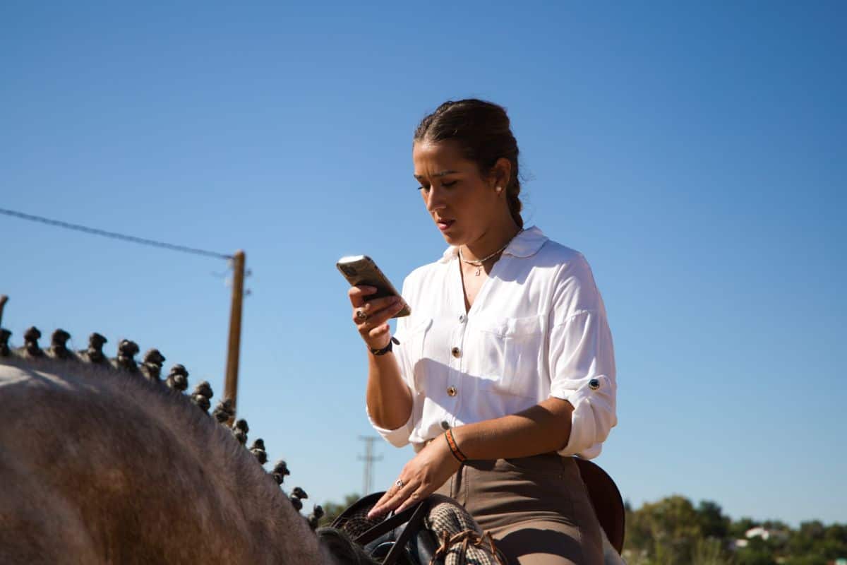 A young woman looking at a phone white sitting on a horse.