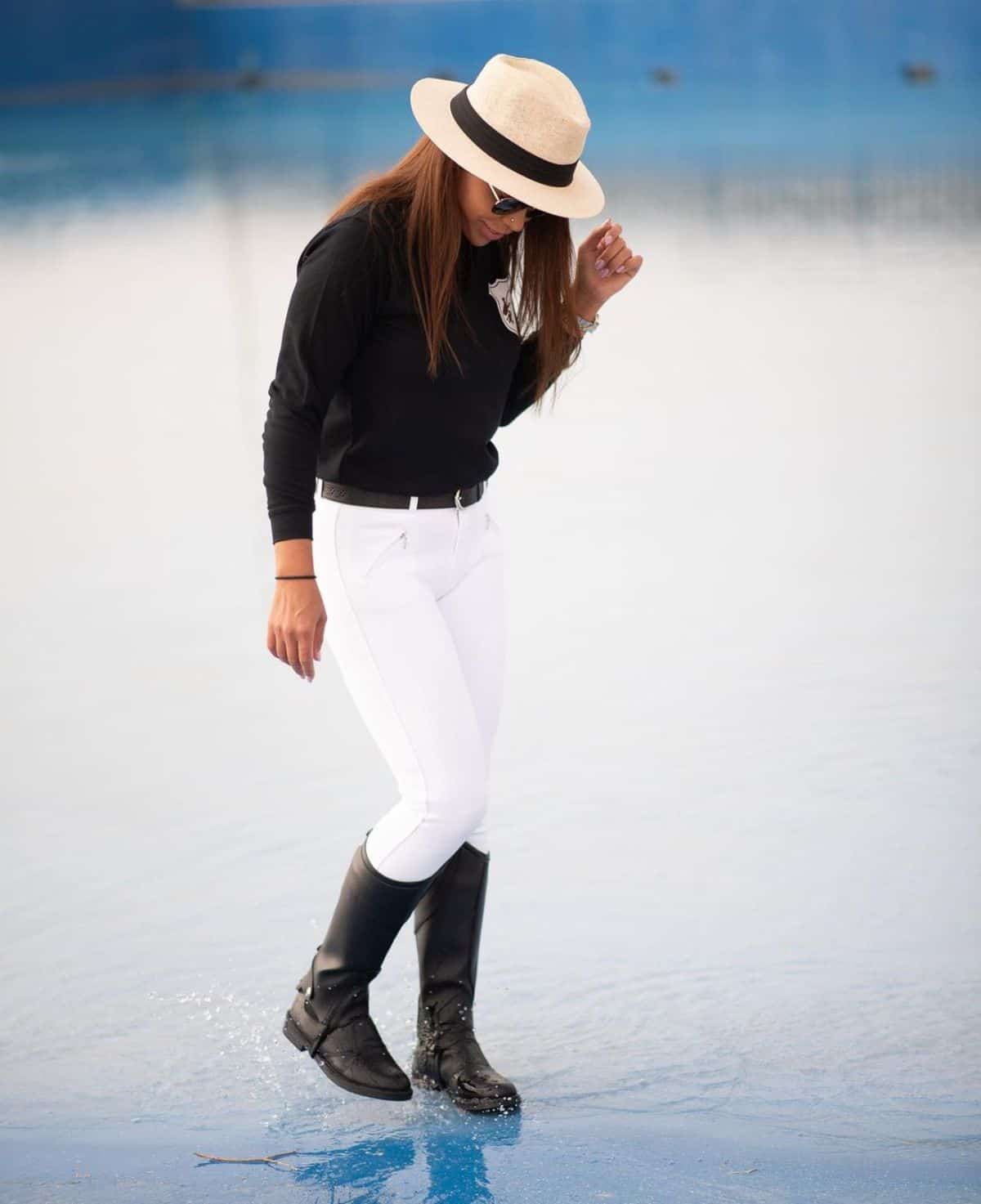 A young woman in an equestrian outfit and a hat stand in shallow water.