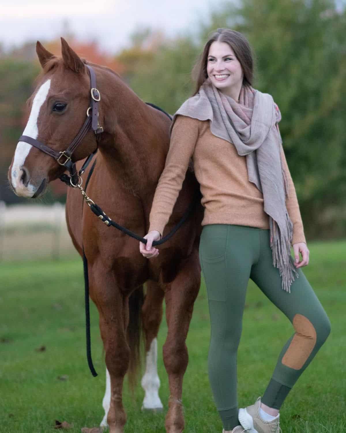A smiling young woman in an equestrian outfit stands next to a brown horse.