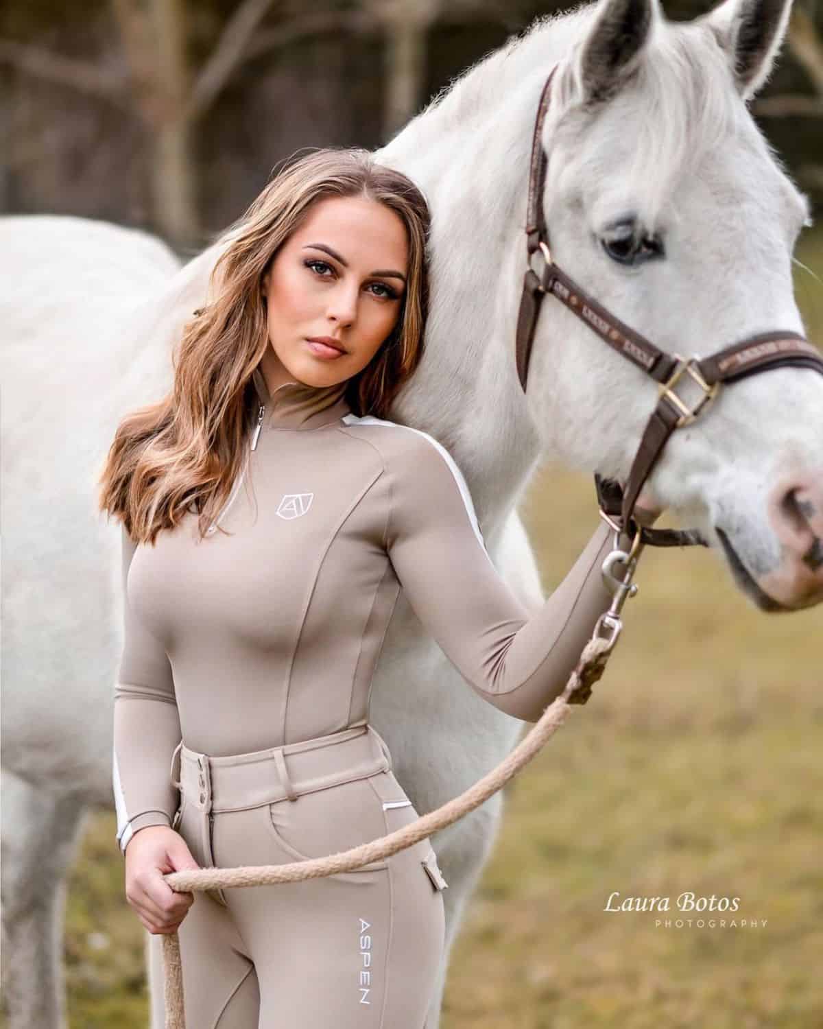 A young woman in an equestrian outfit stands next to a white horse.