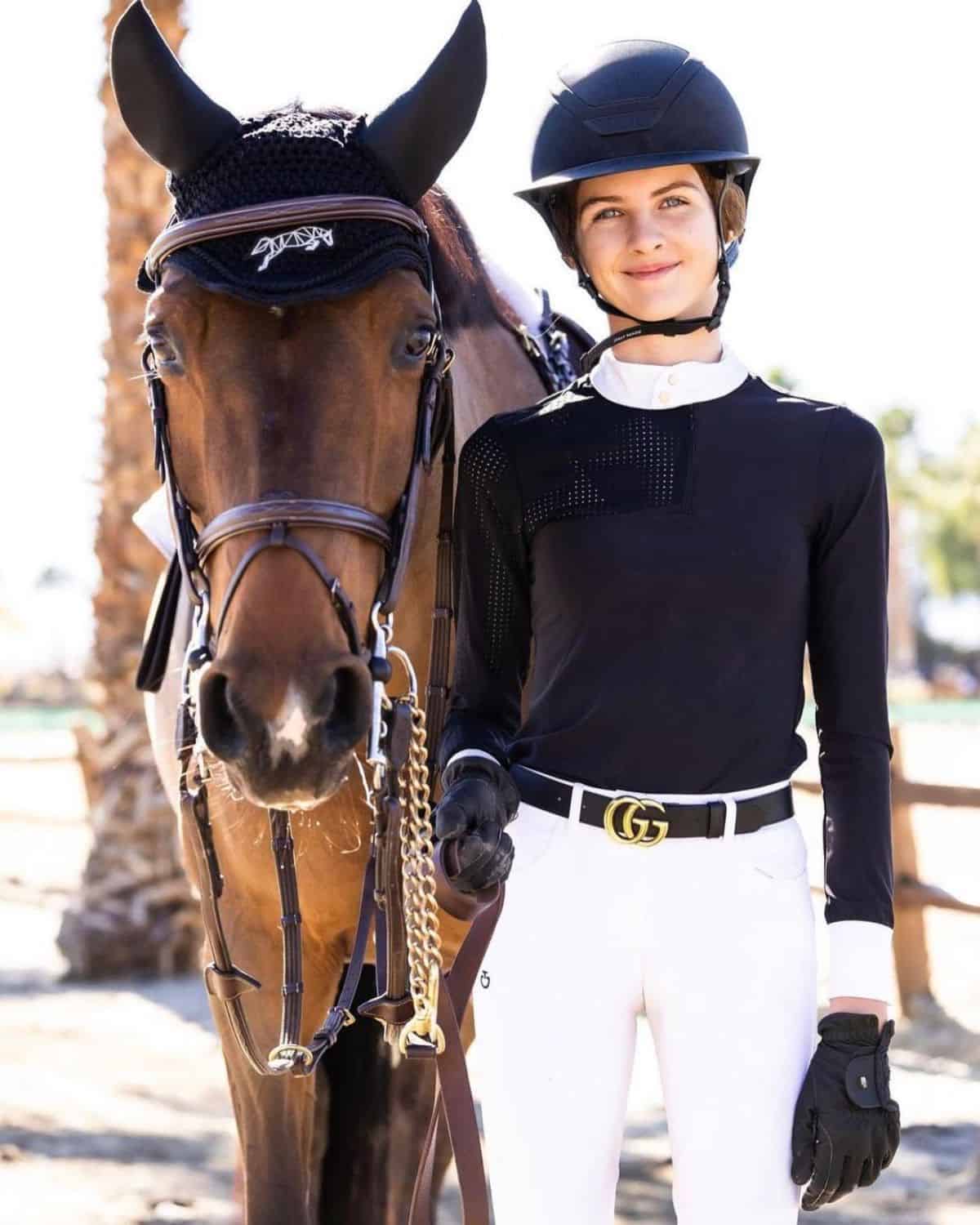 A young woman in an equestrian outfit stands next to a brown horse.