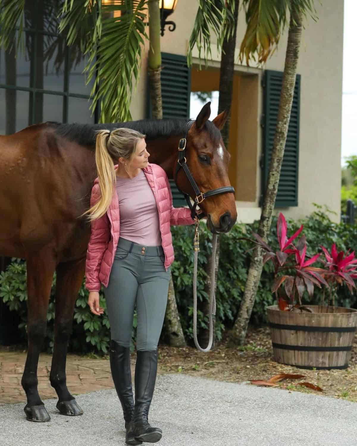 A young woman in an equestrian outfit and a pink jacket stands next to a brown horse.