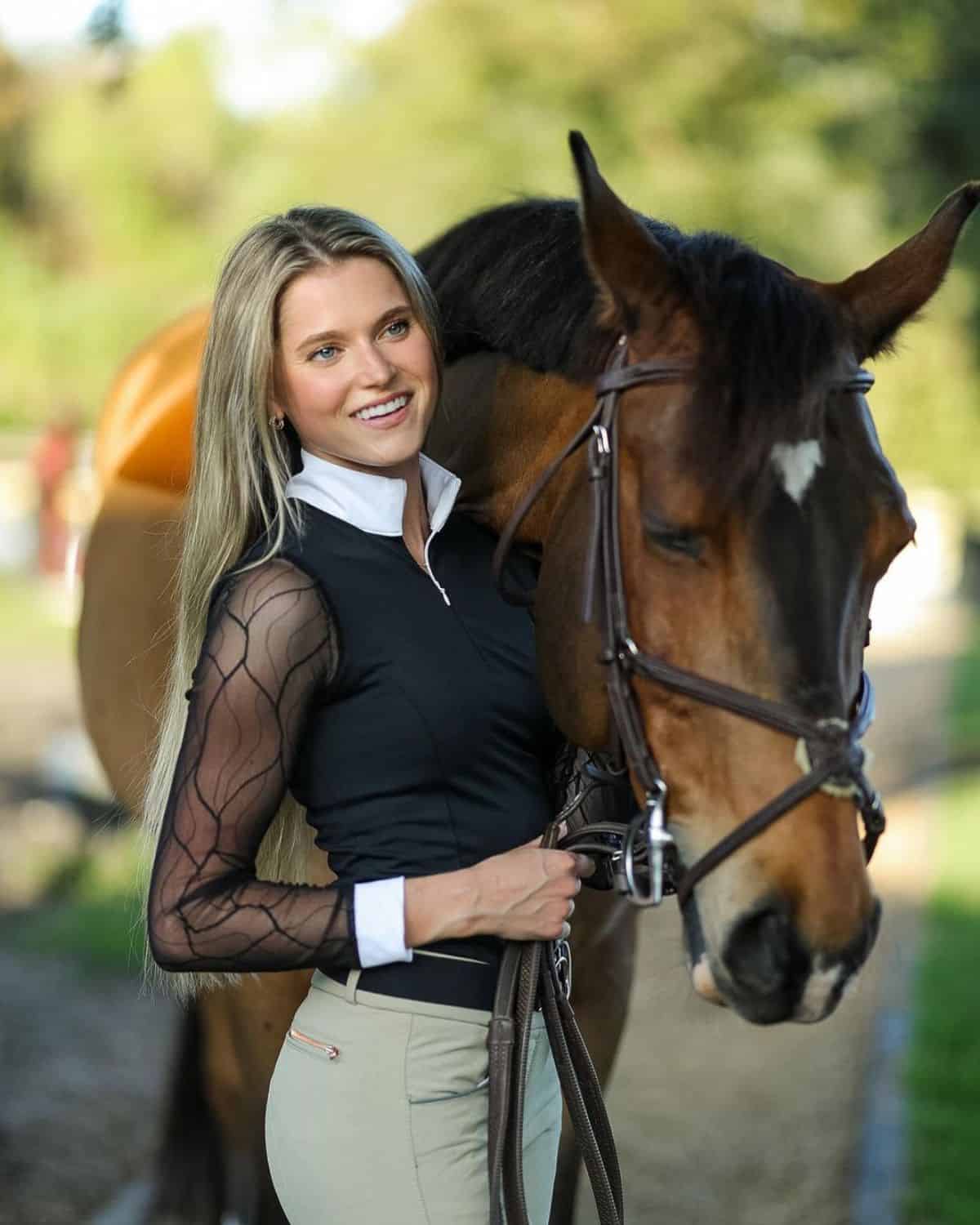 A young woman in an equestrian outfit stands near a brown horse.