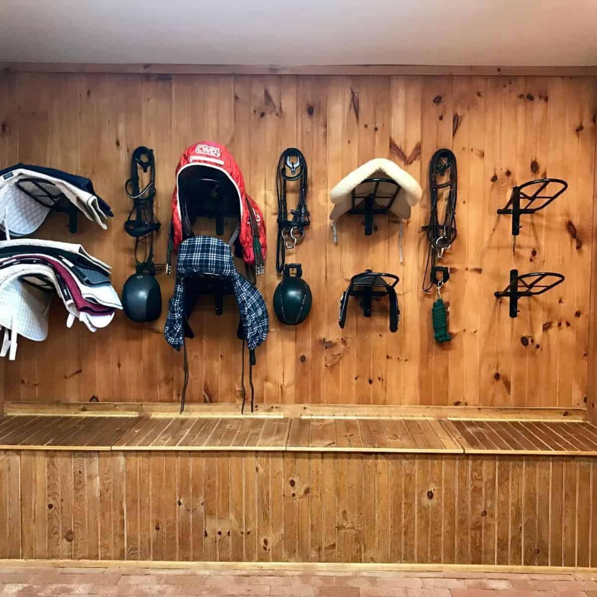 A tack room with halters, helmets, saddles, and bridles hangs on a wooden wall.