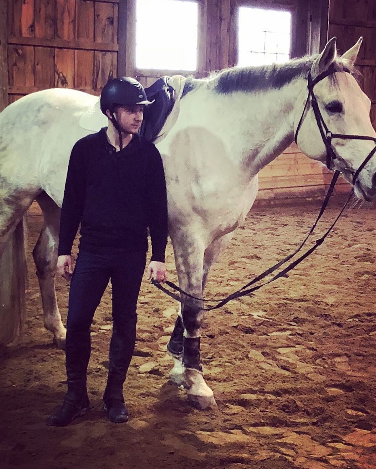 A male Equestrian rider wears a black outfit and stans near a white horse.