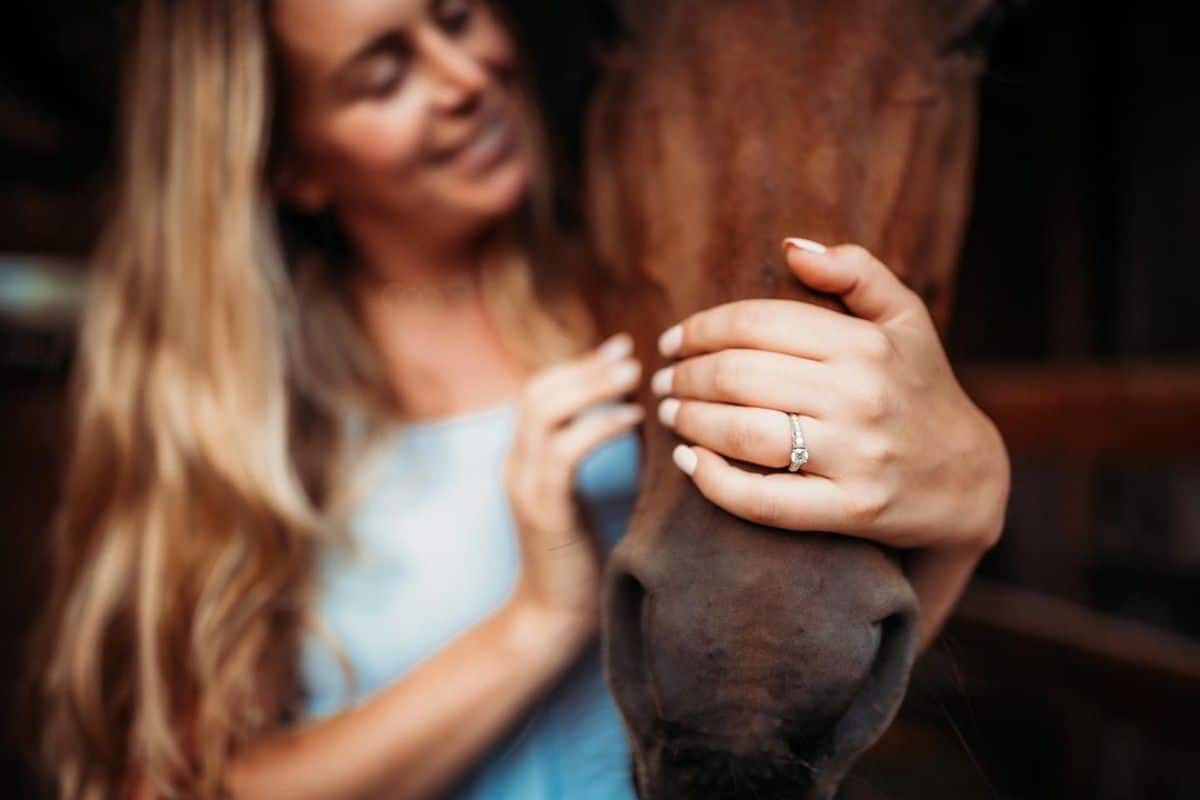 A woman with an engagement ring pets a brown horse.