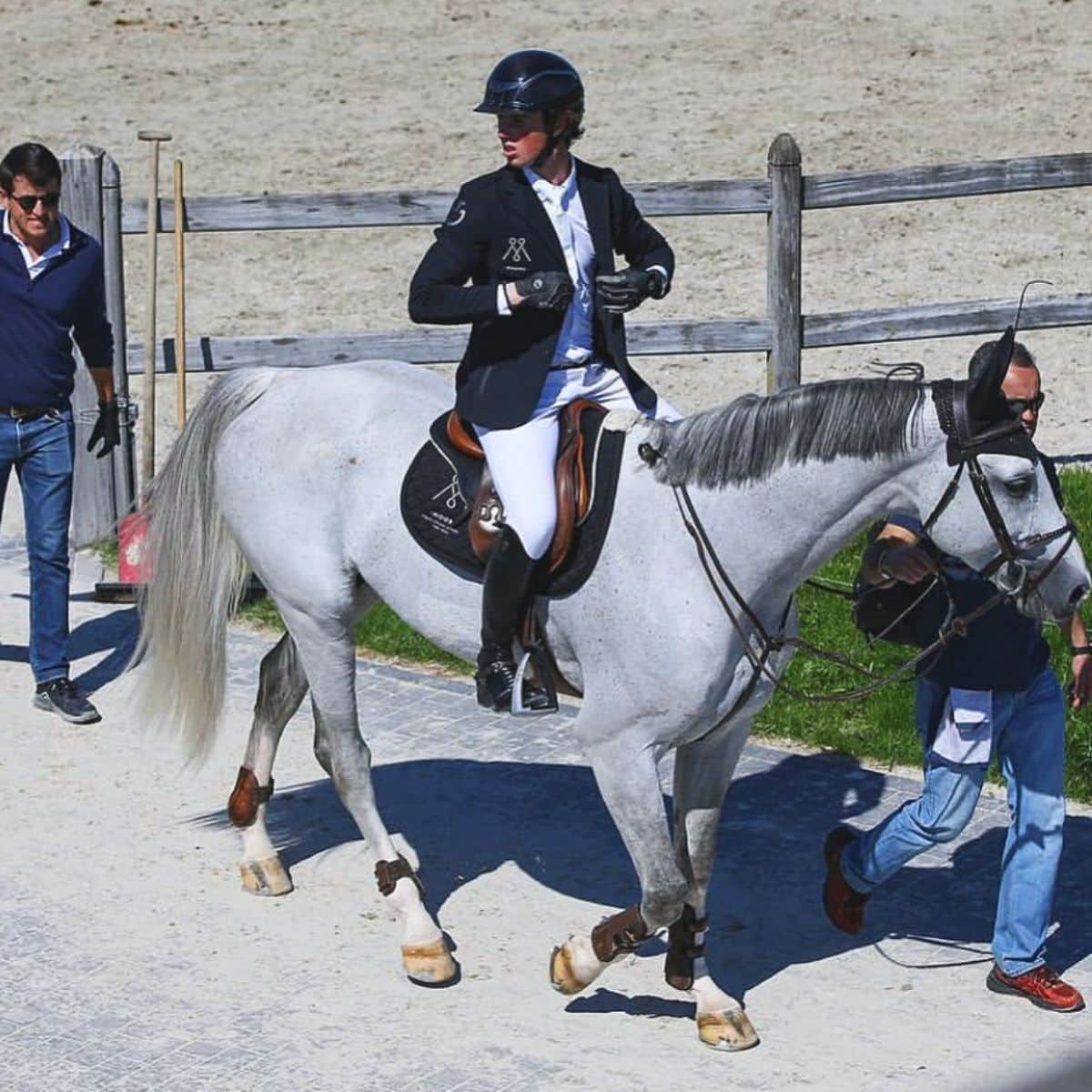 A male Equestrian rider wears a black jaket rides a gray horse.